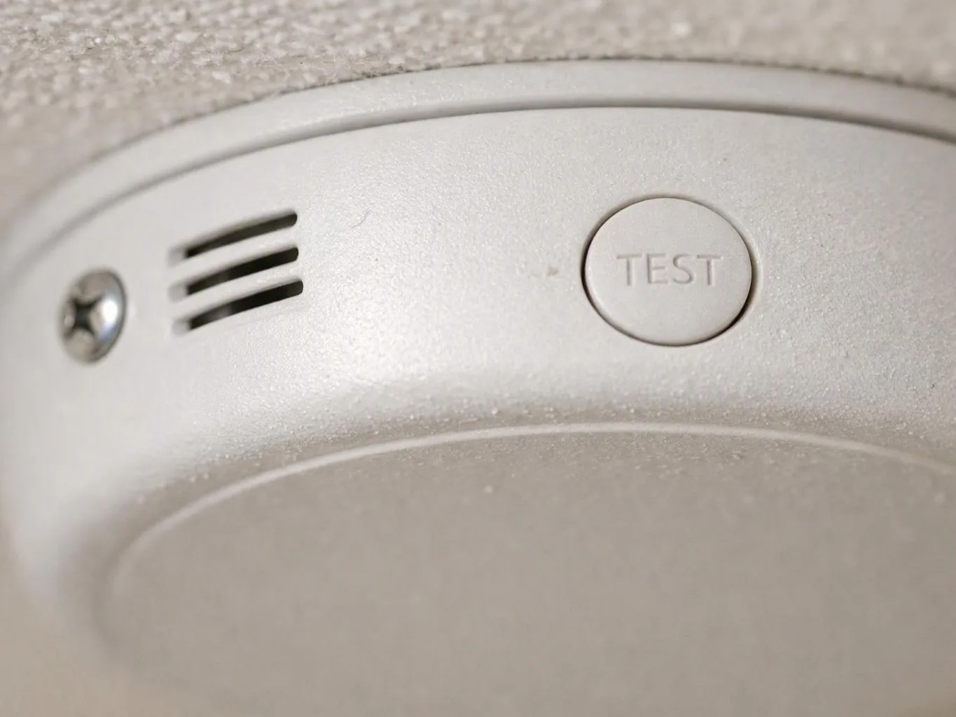 Close-up of a white smoke detector on a ceiling with a TEST button and vent slits
